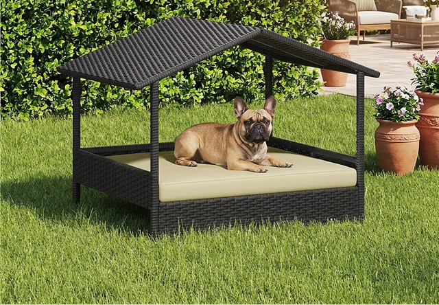 A tan French bulldog lies on a cream cushion inside a black wicker-style house dog bed with a slanted roof, set on a green lawn in a backyard with hedges, patio furniture, and potted flowers.